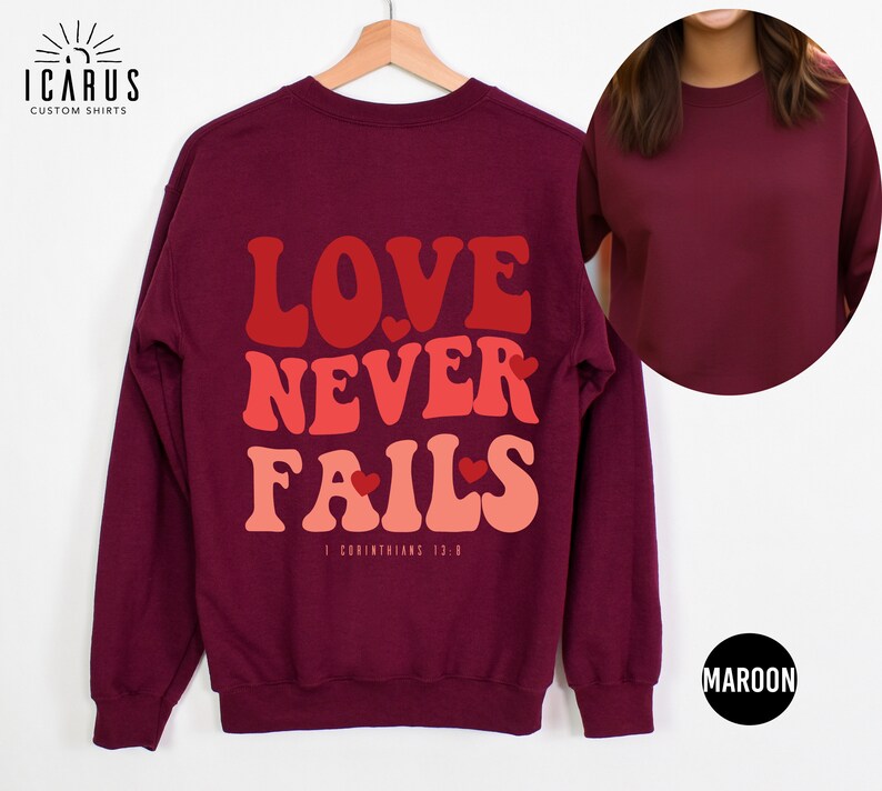 Puede incluir: Sudadera color granate con el texto "LOVE NEVER FAILS" en una fuente retro con corazones. El texto "1 Corintios 13:8" est&aacute; impreso debajo del texto principal.