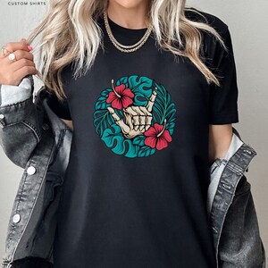 Może przedstawiać: Czarny t-shirt z okrągłym wzorem przedstawiającym szkielet dłoni wykonujący gest rock and rolla, otoczony tropikalnymi liśćmi i czerwonymi kwiatami hibiskusa. Koszulka ma tekst "ICARUS CUSTOM SHIRTS" w lewym g&oacute;rnym rogu.