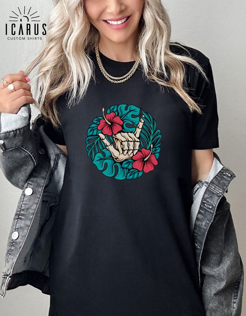 Może przedstawiać: Czarny t-shirt z okrągłym wzorem przedstawiającym szkielet dłoni wykonujący gest rock and rolla, otoczony tropikalnymi liśćmi i czerwonymi kwiatami hibiskusa. Koszulka ma tekst "ICARUS CUSTOM SHIRTS" w lewym g&oacute;rnym rogu.