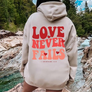Puede incluir: Una sudadera con capucha beige con un gr&aacute;fico rojo, rosa y blanco que dice "LOVE NEVER FAILS" con corazones y el texto "1 Corintios 13:8".