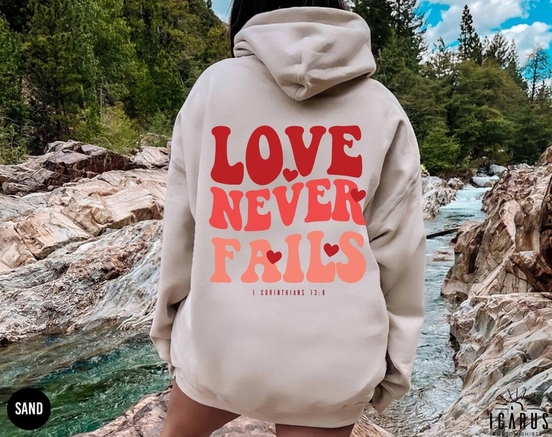 Puede incluir: Una sudadera con capucha beige con un gr&aacute;fico rojo, rosa y blanco que dice "LOVE NEVER FAILS" con corazones y el texto "1 Corintios 13:8".