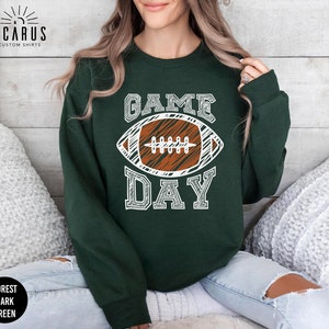 以下が含まれることがあります： 「GAME DAY」の文字と、白と茶色のフットボールのグラフィックがプリントされた、フォレストダークグリーンのスウェットシャツ。クルーネックの長袖スタイルです。「ICARUS CUSTOM SHIRTS」のブランドが左上にプリントされています。