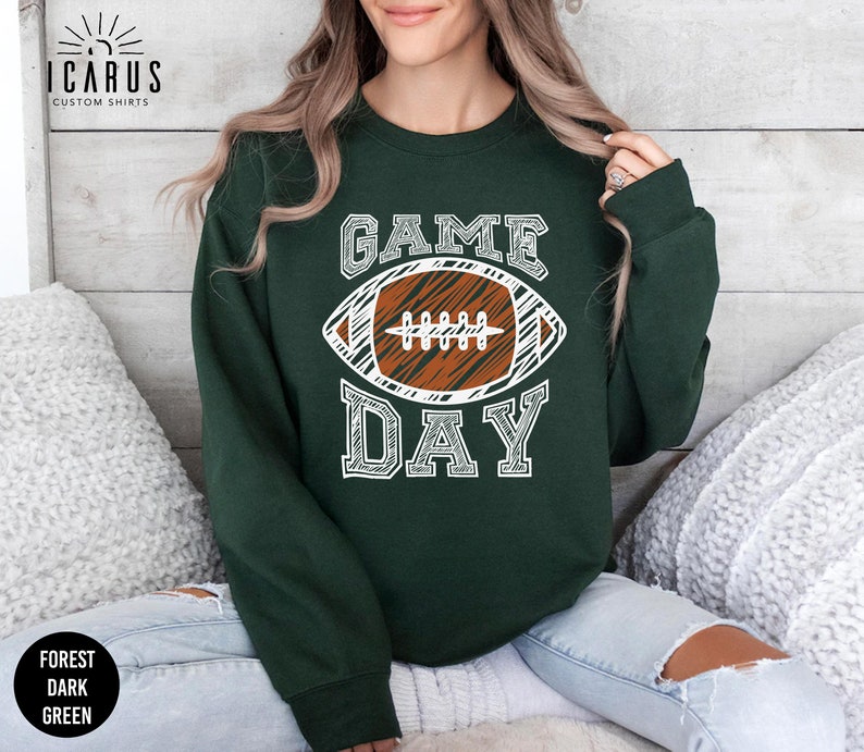 以下が含まれることがあります： 「GAME DAY」の文字と、白と茶色のフットボールのグラフィックがプリントされた、フォレストダークグリーンのスウェットシャツ。クルーネックの長袖スタイルです。「ICARUS CUSTOM SHIRTS」のブランドが左上にプリントされています。