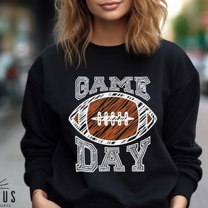 以下が含まれることがあります： 黒いスウェットシャツ。白い文字で「GAME DAY」と書かれており、茶色と白のアメリカンフットボールのグラフィックが描かれています。スウェットシャツはクルーネックと長袖です。「ICARUS CUSTOM SHIRTS」のブランドが下部に印刷されています。