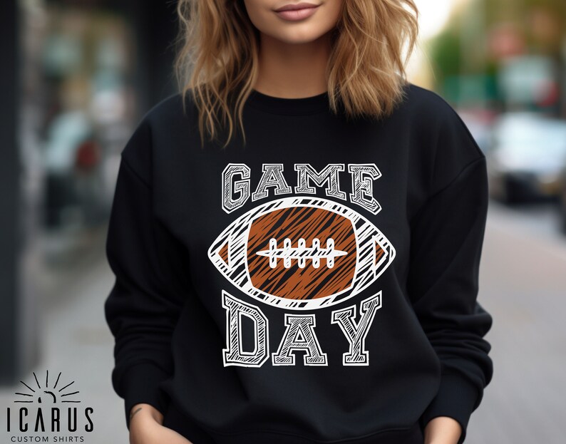 以下が含まれることがあります： 黒いスウェットシャツ。白い文字で「GAME DAY」と書かれており、茶色と白のアメリカンフットボールのグラフィックが描かれています。スウェットシャツはクルーネックと長袖です。「ICARUS CUSTOM SHIRTS」のブランドが下部に印刷されています。