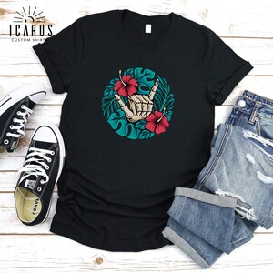 Może przedstawiać: Czarny t-shirt z okrągłym wzorem przedstawiającym szkielet dłoni wykonujący znak shaka, otoczony turkusowymi liśćmi i czerwonymi kwiatami hibiskusa. Koszulka jest pokazana z niebieskimi szortami dżinsowymi i czarnymi trampkami.