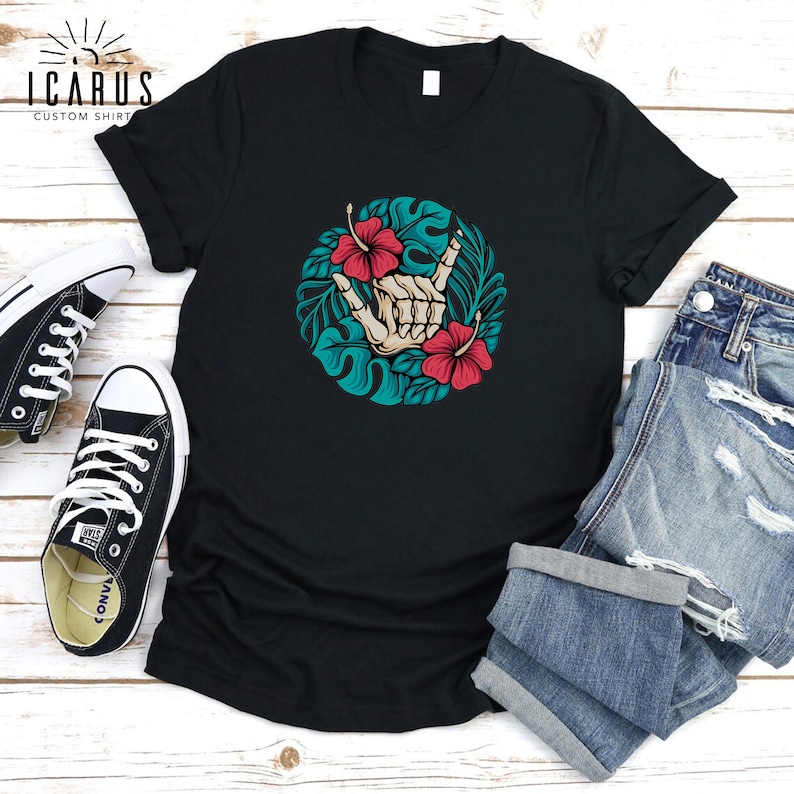 Może przedstawiać: Czarny t-shirt z okrągłym wzorem przedstawiającym szkielet dłoni wykonujący znak shaka, otoczony turkusowymi liśćmi i czerwonymi kwiatami hibiskusa. Koszulka jest pokazana z niebieskimi szortami dżinsowymi i czarnymi trampkami.