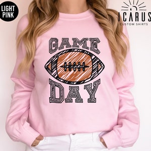 以下が含まれることがあります： ライトピンクのスウェットシャツ。アメフトのグラフィックと「GAME DAY」の文字が黒でプリントされています。アメフトボールは茶色で黒いディテール。スウェットシャツは長袖でクルーネックです。「ICARUS CUSTOM SHIRTS」のブランド名が黒でプリントされています。