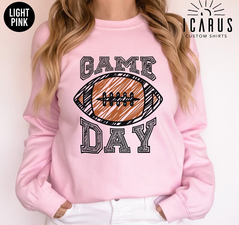 以下が含まれることがあります： ライトピンクのスウェットシャツ。アメフトのグラフィックと「GAME DAY」の文字が黒でプリントされています。アメフトボールは茶色で黒いディテール。スウェットシャツは長袖でクルーネックです。「ICARUS CUSTOM SHIRTS」のブランド名が黒でプリントされています。
