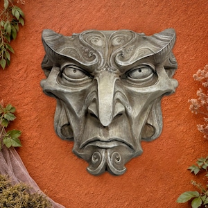 May include: A gray, stone-like wall sculpture of a grotesque face with horns and a stern expression. The sculpture has detailed features, including a prominent nose, furrowed brow, and stylized facial hair. It is mounted on a textured, reddish-orange wall.