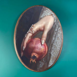 May include: Oval-shaped painting featuring a hand holding a pomegranate. The hand is adorned with a pearl bracelet and has painted nails. The pomegranate is red with a dark, textured base. The painting has a gold frame and a teal background.