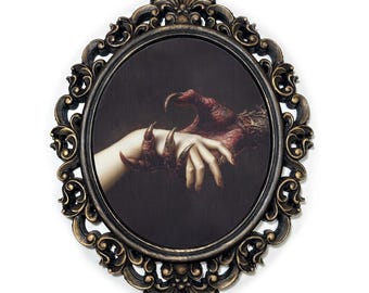 May include: Oval-shaped wall art with a dark background featuring a human hand and a clawed hand. The frame is ornate, with a black and gold finish. The artwork evokes a gothic or horror theme.