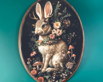 May include: A whimsical illustration of a brown rabbit with antlers wearing a floral crown. The rabbit is surrounded by flowers and mushrooms in a dark forest setting. The illustration is framed in a gold oval.