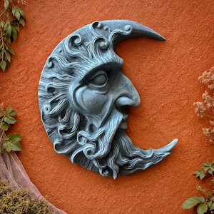 May include: A crescent moon wall sculpture with a weathered, gray finish. The moon features a detailed face with a long beard and flowing hair. The sculpture is mounted on an orange wall, with greenery in the background.