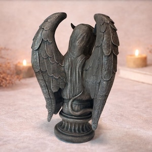 Op de afbeelding: Een grijze beeld van een gevleugelde figuur met een gehoornde kop en een slangestaart. Het beeld heeft grote, gedetailleerde vleugels en staat op een decoratieve basis. De achtergrond bevat brandende kaarsen en een boek, wat een mystieke sfeer cre&euml;ert.
