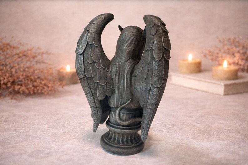Op de afbeelding: Een grijze beeld van een gevleugelde figuur met een gehoornde kop en een slangestaart. Het beeld heeft grote, gedetailleerde vleugels en staat op een decoratieve basis. De achtergrond bevat brandende kaarsen en een boek, wat een mystieke sfeer cre&euml;ert.