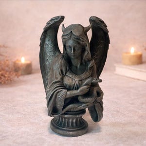 Op de afbeelding: Donkergrijze beeld van een gevleugelde figuur met hoorns, die een slang vasthoudt. Het beeld heeft gedetailleerde vleugels en is gemonteerd op een decoratieve basis. De figuur draagt een gedetailleerde jurk.