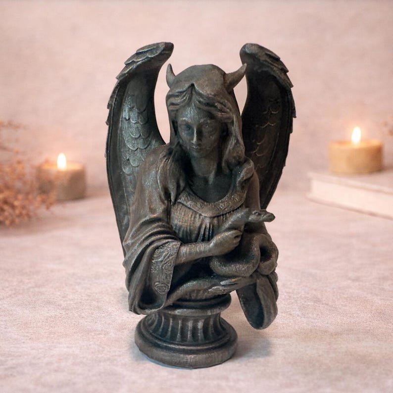 Op de afbeelding: Donkergrijze beeld van een gevleugelde figuur met hoorns, die een slang vasthoudt. Het beeld heeft gedetailleerde vleugels en is gemonteerd op een decoratieve basis. De figuur draagt een gedetailleerde jurk.