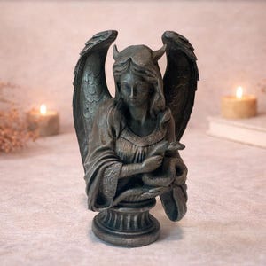 Op de afbeelding: Een bronzen beeld van een gevleugelde figuur met hoorns, die een slang vasthoudt. Het beeld heeft gedetailleerde vleugels en staat op een decoratieve sokkel. De achtergrond bevat brandende kaarsen en een boek, wat een mystieke sfeer cre&euml;ert.