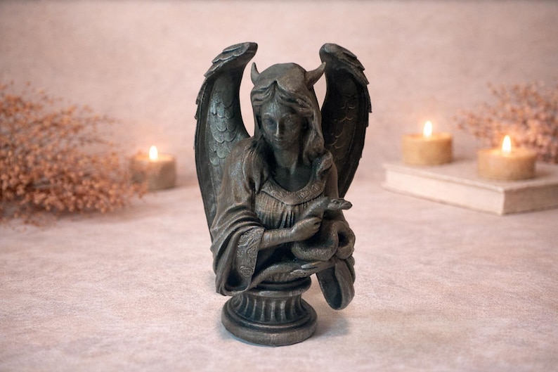Op de afbeelding: Een bronzen beeld van een gevleugelde figuur met hoorns, die een slang vasthoudt. Het beeld heeft gedetailleerde vleugels en staat op een decoratieve sokkel. De achtergrond bevat brandende kaarsen en een boek, wat een mystieke sfeer cre&euml;ert.