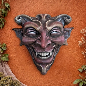 May include: A detailed, wall-mounted gargoyle face sculpture. The sculpture features a sinister grin with sharp teeth, a prominent nose, and curled horns. The color palette includes shades of purple, red, and gray, with white highlights on the eyes.
