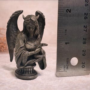 Op de afbeelding: Een gedetailleerd beeld in antieke stijl van een gevleugelde figuur met hoorns, die een slang vasthoudt. Het beeld is ongeveer 5 cm hoog en is gemaakt van een donker, metalen materiaal. De figuur zit op een decoratieve basis.