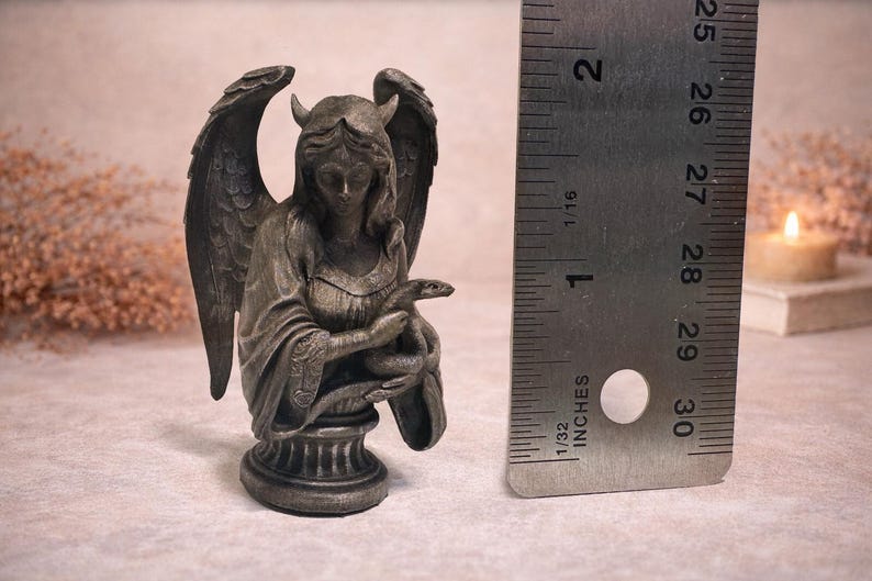 Op de afbeelding: Een gedetailleerd beeld in antieke stijl van een gevleugelde figuur met hoorns, die een slang vasthoudt. Het beeld is ongeveer 5 cm hoog en is gemaakt van een donker, metalen materiaal. De figuur zit op een decoratieve basis.