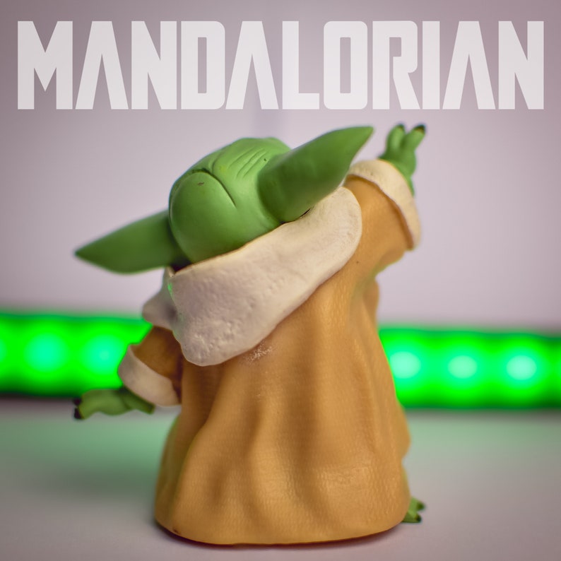 K&ouml;nnte beinhalten: Eine kleine gr&uuml;ne und braune Figur eines Charakters aus dem Star Wars-Franchise, bekannt als "The Child" oder "Baby Yoda", tr&auml;gt eine braune Robe mit einem wei&szlig;en Pelzkragen. Die Figur steht auf einer wei&szlig;en Oberfl&auml;che mit einem gr&uuml;nen Lichtstreifen im Hintergrund. Der Text "MANDALORIAN" ist oben im Bild sichtbar.