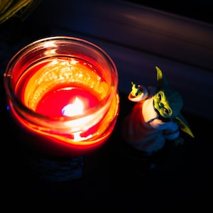 K&ouml;nnte beinhalten: Eine rote Kerze in einem Glasgef&auml;&szlig; ist angez&uuml;ndet und leuchtet. Im Hintergrund befindet sich eine gr&uuml;n-braune Figur.