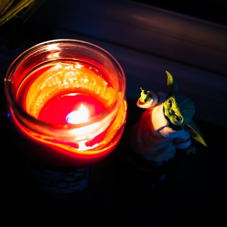 K&ouml;nnte beinhalten: Eine rote Kerze in einem Glasgef&auml;&szlig; ist angez&uuml;ndet und leuchtet. Im Hintergrund befindet sich eine gr&uuml;n-braune Figur.
