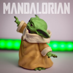 K&ouml;nnte beinhalten: Eine gr&uuml;ne und braune Spielzeugfigur eines Charakters aus dem Star Wars-Franchise, bekannt als "The Child" oder "Baby Yoda", tr&auml;gt eine braune Robe mit einem wei&szlig;en Pelzkragen. Die Figur steht auf einer wei&szlig;en Oberfl&auml;che mit einem gr&uuml;nen Lichtstreifen im Hintergrund. Der Text "MANDALORIAN" ist &uuml;ber der Figur sichtbar.