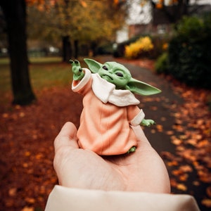 K&ouml;nnte beinhalten: Eine kleine gr&uuml;ne und braune Figur eines Charakters aus dem Star Wars-Franchise, die eine pfirsichfarbene Robe mit wei&szlig;em Kragen tr&auml;gt und in einer ausgestreckten Hand steht.