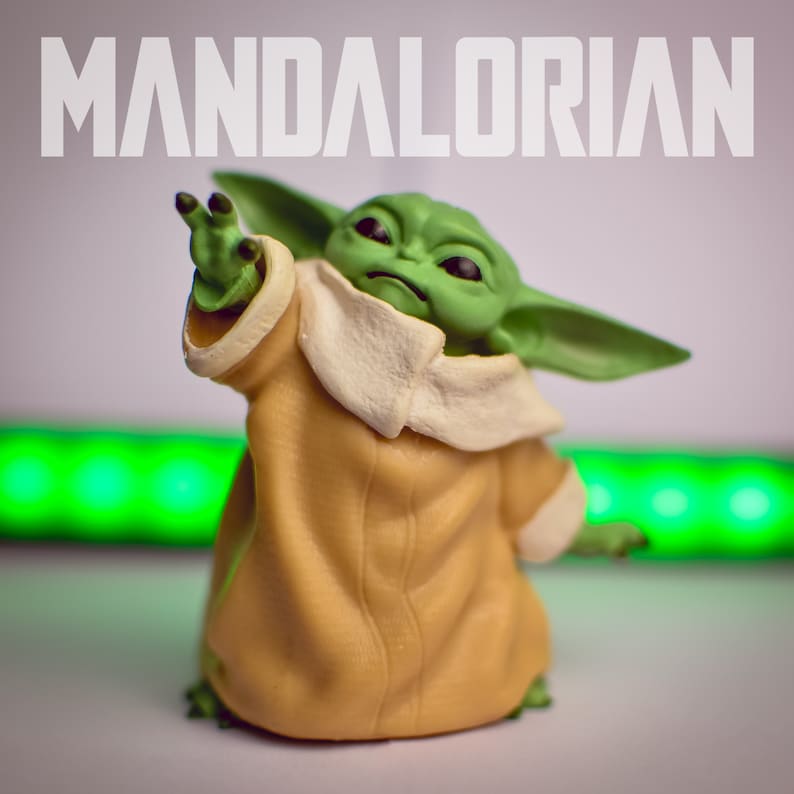 K&ouml;nnte beinhalten: Eine gr&uuml;ne und gelbe Spielzeugfigur eines Charakters aus dem Star Wars-Franchise, bekannt als "The Child" oder "Baby Yoda", mit dem Wort "MANDALORIAN" in wei&szlig;er Schrift &uuml;ber der Figur.