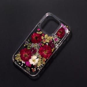 Puede incluir: Funda transparente para teléfono con rosas rojas secas, flores blancas y moradas, y hojas verdes. El diseño transparente de la funda resalta el arreglo floral. Un accesorio protector para teléfono móvil.