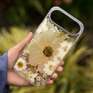 May include: A clear protective phone case featuring an arrangement of pressed white and cream-colored flowers, including a large central bloom, small daisies, and delicate greenery embedded within the transparent material of the cover.
