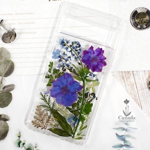 May include: A clear phone case with a pressed flower design. The flowers are purple, blue, and green. The case is made of clear plastic and has a smooth finish.