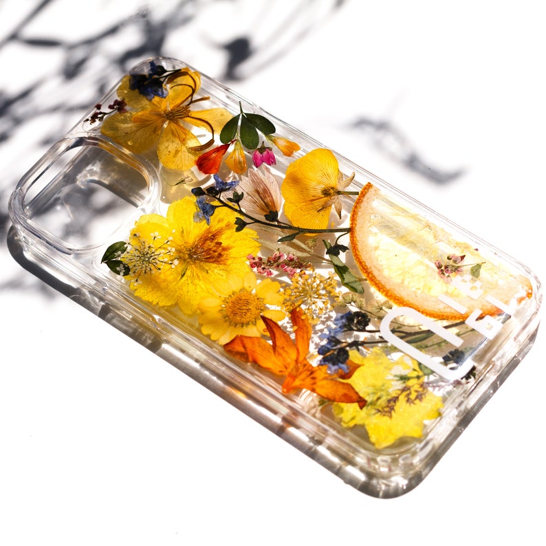 May include: A clear phone case with a pressed flower design. The case features a variety of yellow, orange, and blue flowers, as well as a slice of dried citrus fruit. The case also has the word "Cute" printed on it in white.