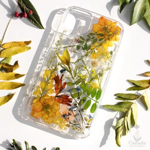 May include: A clear phone case with pressed flowers and leaves. The flowers are yellow, orange, and red. The leaves are green. The case is designed to fit a smartphone.