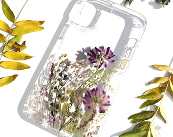 May include: A clear phone case with pressed purple and white flowers and greenery inside. The case is designed for a smartphone with a camera bump.