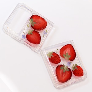 May include: Two clear phone cases with embedded dried strawberries and flowers. The cases are open to show the inside.