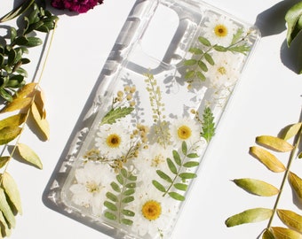May include: A clear phone case with a pressed flower design. The case features white daisies and green leaves pressed between the layers of the case.