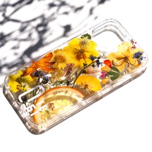 May include: A clear phone case with a pressed flower design. The case features yellow, orange, and red flowers, as well as a slice of lemon. The case has a white text label that says "Life".