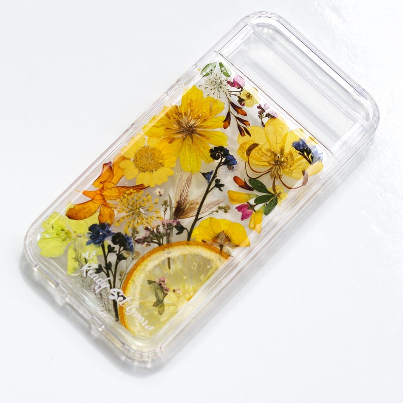 May include: A clear phone case with a pressed flower design. The case features yellow, orange, and white flowers, as well as a slice of lemon. The text "Kindly & Green" is visible in the lower left corner.