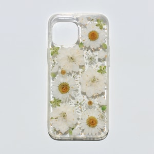May include: A clear phone case with a pressed flower design. The case features white flowers and green leaves with gold glitter accents.