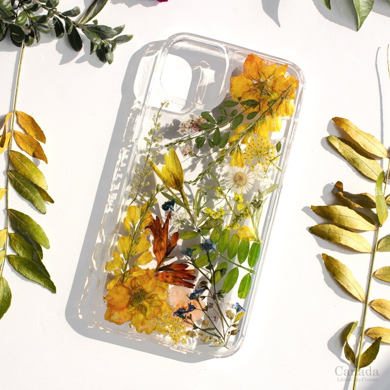 May include: A clear phone case with pressed dried flowers and leaves. The flowers are yellow, orange, and red. The leaves are green. The case is made of clear plastic and has a raised edge around the camera lens.