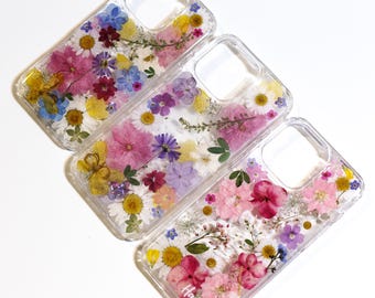 May include: Three clear phone cases with pressed flowers. The cases are decorated with different arrangements of pink, purple, white, and yellow flowers. The cases are for an iPhone 13 Pro Max.