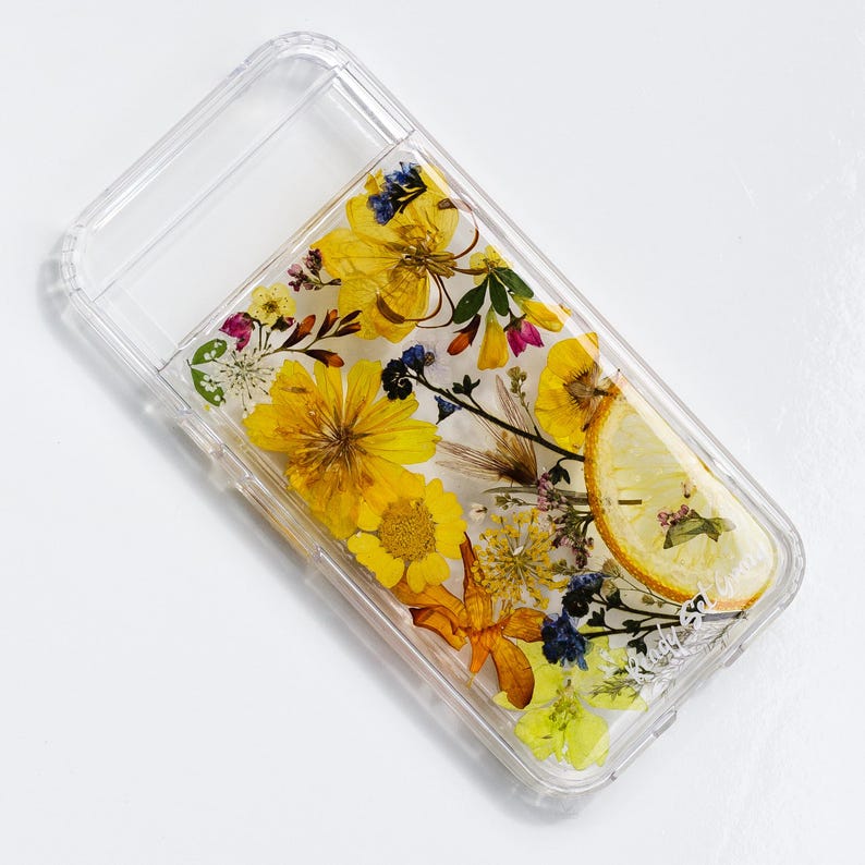 May include: A clear phone case with a pressed flower design. The case features yellow, white, and orange flowers, as well as green leaves. A slice of lemon is also visible in the design.