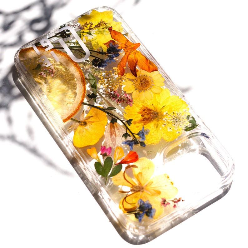 May include: A clear phone case with pressed flowers and a lemon slice. The flowers are yellow, orange, and blue. The case is designed for a smartphone with a camera cutout.