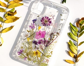 May include: A clear phone case featuring an arrangement of pressed dried flowers in shades of purple, pink, and yellow. The floral design is embedded within the transparent material, showcasing delicate petals, stems, and green leaves throughout.