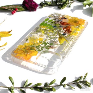 May include: A clear phone case with pressed flowers and leaves. The flowers are yellow, orange, and white. The case is designed for a smartphone with a camera cutout.